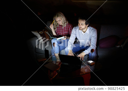 Freelance, education. Man and woman, couple sitting on sofa at home in the evening and studying, working with little light of battery lamp. No electricity, blackout Freelance, education. Man and woman, couple sitting on sofa at home in the evening and studying, working with little light of battery lamp. No electricity, blackout 98825425