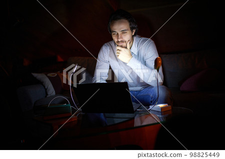 Adjusting to unstable electricity. Man, freelancer sitting on sofa at home in evening and working on laptop with battery lamp light. Blackout, no electricity 98825449