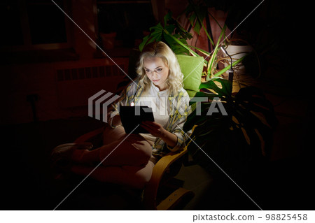 Battery powered lamp light. Young beautiful woman sitting in dark room with laptop at home in the evening. no electricity, blackout Battery powered lamp light. Young beautiful woman sitting in dark room with laptop at home in the evening. no electricity, blackout 98825458
