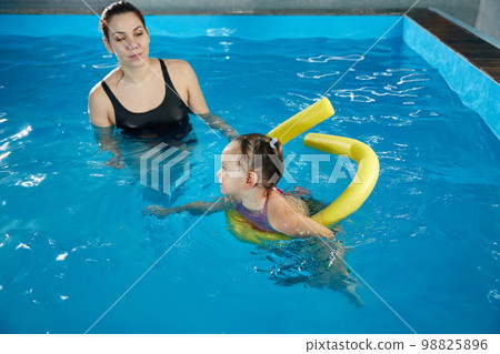 Preschool girllearning to swim in pool with foam noodle with young trainer 98825896