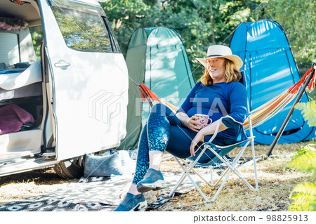 Adult woman relaxing, drinking tea while sitting in the travel chair near camper car vehicle and tents. Weekend trip activity. Female living on camper car and travel the world. Caravan car Vacation Adult woman relaxing, drinking tea while sitting in the travel chair near camper car vehicle and tents. Weekend trip activity. Female living on camper car and travel the world. Caravan car Vacation 98825913