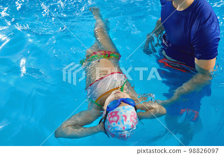 Trainer teaching preschool girl how to swim on her back in indoor pool 98826097