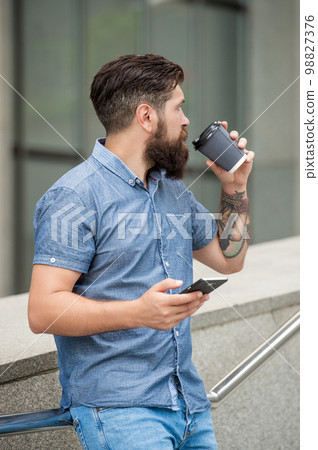 Bearded guy texting via mobile phone while drinking coffee outdoors Chatting online on mobile device Bearded guy texting via mobile phone while drinking coffee outdoors Chatting online on mobile device 98827376