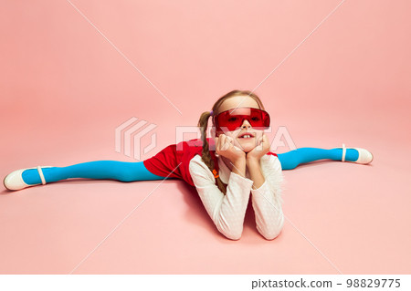 Girl, child in red dress, blue tights and red glasses sitting on twine, posing over pink studio background. Concept of emotions, childhood 98829775