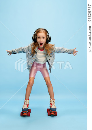 Little beautiful girl, child in shorts and jeans jacket emotionally posing in headphones, moving on rollers over blue studio background. Concept of emotions, childhood Little beautiful girl, child in shorts and jeans jacket emotionally posing in headphones, moving on rollers over blue studio background. Concept of emotions, childhood 98829787
