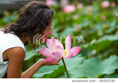 A young, beautiful woman is resting by the lotus pond. Portrait of a beautiful woman in a green park by a lotus pond with blooming lotuses 98830190