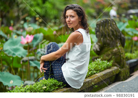 A young, beautiful woman is resting by the lotus pond. Portrait of a beautiful woman in a green park by a lotus pond with blooming lotuses 98830191