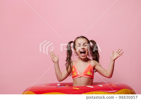 Mischievous little girl in swimwear, making faces, grimacing, posing with pink swimming ring donut, isolated over pink color background, copy ad space. Summer holidays and beach entertainment concept 98830857