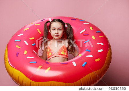Beautiful Caucasian little child girl with two ponytails, wearing a bright stylish swimsuit, smiles looking at camera, posing with a big inflatable donut swimming ring, isolated over pink background Beautiful Caucasian little child girl with two ponytails, wearing a bright stylish swimsuit, smiles looking at camera, posing with a big inflatable donut swimming ring, isolated over pink background 98830860