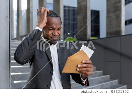 Upset African American man standing near the courthouse, holding an envelope with a letter in his hands. He holds his head, Received documents for divorce, financial agreement, alimony, lawsuit. Upset African American man standing near the courthouse, holding an envelope with a letter in his hands. He holds his head, Received documents for divorce, financial agreement, alimony, lawsuit. 98831502