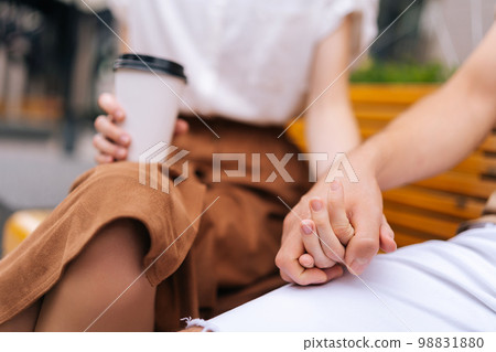 Close-up cropped shot of unrecognizable happy young couple in love holding hands sitting on bench and drinking takeaway coffee enjoying time together outdoors on summer day, blurred background. Close-up cropped shot of unrecognizable happy young couple in love holding hands sitting on bench and drinking takeaway coffee enjoying time together outdoors on summer day, blurred background. 98831880