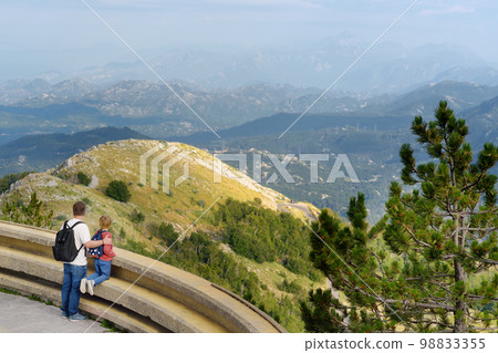 Preteen boy and father admiring of amazing aerial view of range near Kotor Bay, Montenegro. Breathing tonal perspective panorama of Balkan mountains taking from Lovchen National Park. 98833355