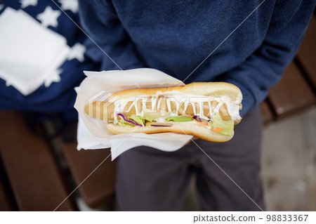 Little boy eating hot dog in public park. Child enjoying his to go meal outside. Fast food is a junk food. Overweight kids. 98833367