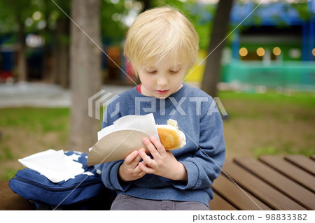 Little boy eating hot dog in public park. Child enjoying his to go meal outside. Fast food is a junk food. Overweight kids. 98833382