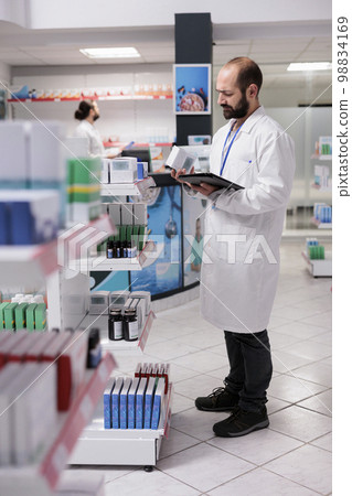 Pharmacist looking at pills packages typing inventory information on tablet computer in drugstore. Pharmacy is a trusted source for high-quality and effective medication and treatment options. Pharmacist looking at pills packages typing inventory information on tablet computer in drugstore. Pharmacy is a trusted source for high-quality and effective medication and treatment options. 98834169