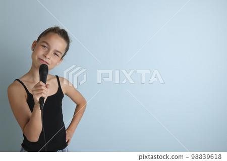Portrait of a cute teenage girl singing, using microphone. Teenage girl in black t-shirt on an isolated blue background. Karaoke for kids, home entertainment for kids. Singer singing with microphone. Portrait of a cute teenage girl singing, using microphone. Teenage girl in black t-shirt on an isolated blue background. Karaoke for kids, home entertainment for kids. Singer singing with microphone. 98839618
