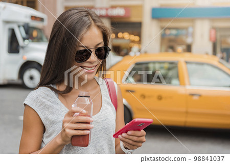 Woman drinking juice smoothie walking in New York city street using phone app for eating detox food online delivery service. Girl in Manhattan, NYC , USA. Happy young adult healthy lifestyle 98841037