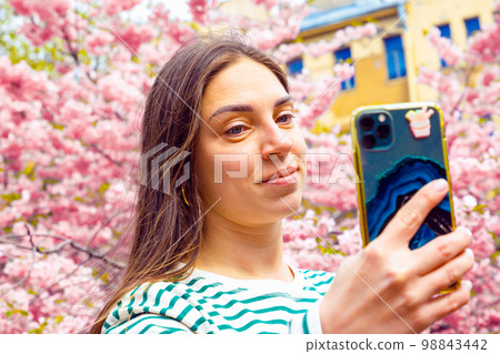 Cheerful young woman talking her picture with her cell phone. Caucasian female model photographing herself with smart phone at spring park. Cheerful young woman talking her picture with her cell phone. Caucasian female model photographing herself with smart phone at spring park. 98843442