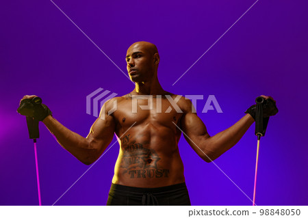 Sporty man performs exercises with expander on violet studio background. Strength and motivation Sporty man performs exercises with expander on violet studio background. Strength and motivation 98848050