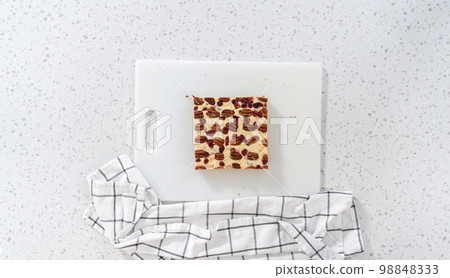 Flat lay. Cutting white chocolate cranberry pecan fudge into small pieces on a white cutting board. 98848333