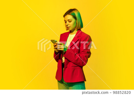 Half-length portrait of young emotional girl with blonde hair wearing red jacket using phone over yellow background. Concept of youth, student college life, business and education 98852820