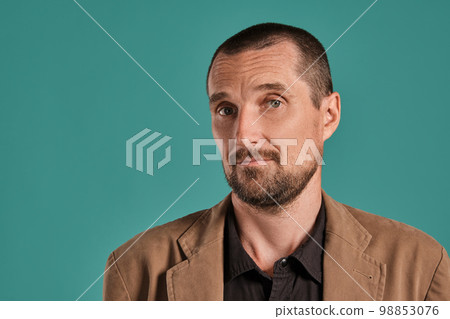 Middle-aged man with beard and mustache, wears black shirt and brown jacket posing against a blue background. Sincere emotions concept. Middle-aged man with beard and mustache, wears black shirt and brown jacket posing against a blue background. Sincere emotions concept. 98853076