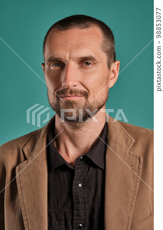 Middle-aged man with beard and mustache, wears black shirt and brown jacket posing against a blue background. Sincere emotions concept. 98853077