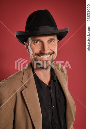 Middle-aged man with beard and mustache, wears black hat and brown jacket posing against a red background. Sincere emotions concept. Middle-aged man with beard and mustache, wears black hat and brown jacket posing against a red background. Sincere emotions concept. 98853090