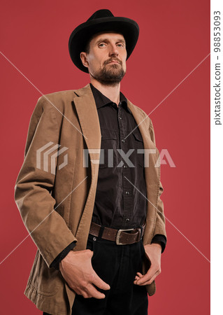 Middle-aged man with beard and mustache, wears black hat and brown jacket posing against a red background. Sincere emotions concept. Middle-aged man with beard and mustache, wears black hat and brown jacket posing against a red background. Sincere emotions concept. 98853093
