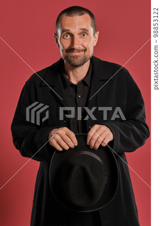 Middle-aged man with beard and mustache, wears black hat and jacket posing against a red background. Sincere emotions concept. 98853122