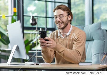 Young handsome man businessman, freelancer sits in the office at the computer and uses the phone. Type messages, chstting,, read news, smile. 98859716