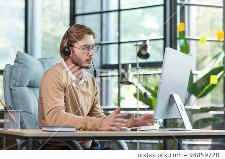 Online psychologist. A young man, a psychologist, a psychotherapist sits in the office in a headset, works at a computer, consults, talks via video call to clients, patients. Online psychologist. A young man, a psychologist, a psychotherapist sits in the office in a headset, works at a computer, consults, talks via video call to clients, patients. 98859720