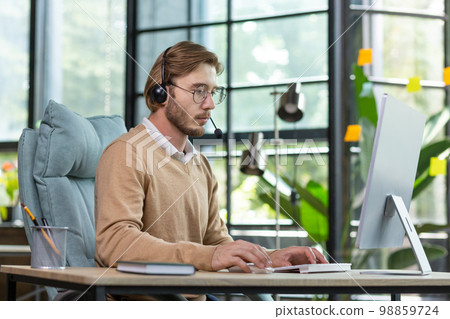 Online trade, support, consultation. Portrait of a young handsome man who sits in the office at the table in a headset, concentrates on working at the computer. Online trade, support, consultation. Portrait of a young handsome man who sits in the office at the table in a headset, concentrates on working at the computer. 98859724