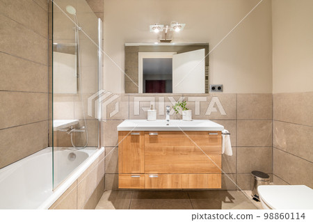 Bathroom in beautiful beige color. Vanity top sink with wooden drawers. Bathtub is fenced with protective glass from splashes. Front door is reflected in square large mirror opposite. Bathroom in beautiful beige color. Vanity top sink with wooden drawers. Bathtub is fenced with protective glass from splashes. Front door is reflected in square large mirror opposite. 98860114