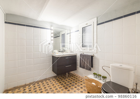 Large bathroom with glossy white walls and beige marble mosaic floor. In the corner is a sink on a vanity with black wooden drawers and a mirror with bright lamps. Near a window for ventilation. Large bathroom with glossy white walls and beige marble mosaic floor. In the corner is a sink on a vanity with black wooden drawers and a mirror with bright lamps. Near a window for ventilation. 98860116