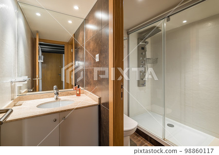 Spacious bathroom with separate toilet area and an electronic shower cabin and washbasin on vanity with large full-wall mirror reflecting entrance door to bathroom. Chocolate colored marble walls. Spacious bathroom with separate toilet area and an electronic shower cabin and washbasin on vanity with large full-wall mirror reflecting entrance door to bathroom. Chocolate colored marble walls. 98860117