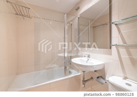 Spacious bathroom with soft peach walls, small sink and square mirror. Next to it is white toilet bowl and glass shelves above it for various accessories. On other side is white, clean bathtub. Spacious bathroom with soft peach walls, small sink and square mirror. Next to it is white toilet bowl and glass shelves above it for various accessories. On other side is white, clean bathtub. 98860128