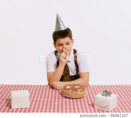 At birthday party. Cute little boy sitting at table covered plaid table cloth and looking at cake isolated on white background. Bored. Concept of emotions, retro style, fashion, family. 98861167