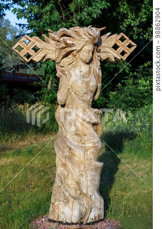A wooden sculpture of a dancing girl stands against a blurred background of a green park. 06.19.2022. Kyiv. Ukraine. 98862904