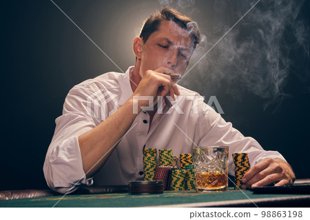 Handsome emotional man is playing poker sitting at the table in casino against a white spotlight. Handsome emotional man is playing poker sitting at the table in casino against a white spotlight. 98863198