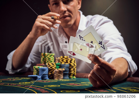Handsome emotional man is playing poker sitting at the table in casino against a white spotlight. Handsome emotional man is playing poker sitting at the table in casino against a white spotlight. 98863206