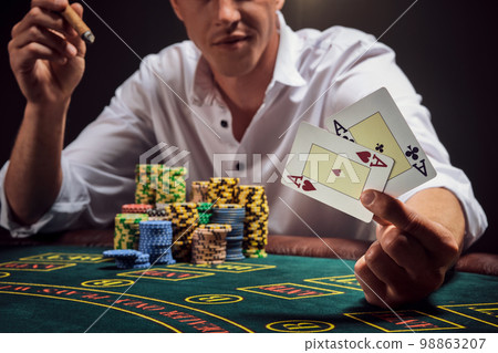 Handsome emotional man is playing poker sitting at the table in casino against a white spotlight. Handsome emotional man is playing poker sitting at the table in casino against a white spotlight. 98863207