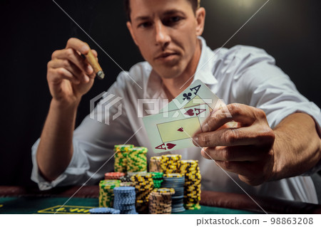 Handsome emotional man is playing poker sitting at the table in casino against a white spotlight. Handsome emotional man is playing poker sitting at the table in casino against a white spotlight. 98863208