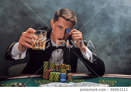 Handsome emotional man is playing poker sitting at the table in casino against a white spotlight. Handsome emotional man is playing poker sitting at the table in casino against a white spotlight. 98863211