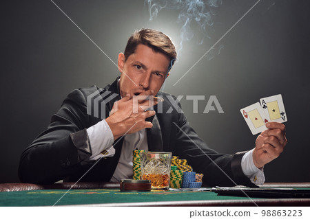 Handsome emotional man is playing poker sitting at the table in casino against a white spotlight. Handsome emotional man is playing poker sitting at the table in casino against a white spotlight. 98863223