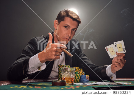 Handsome emotional man is playing poker sitting at the table in casino against a white spotlight. Handsome emotional man is playing poker sitting at the table in casino against a white spotlight. 98863224