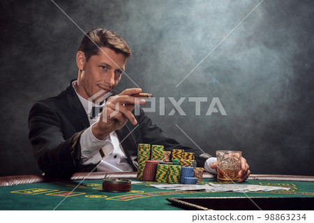 Handsome emotional man is playing poker sitting at the table in casino against a white spotlight. 98863234