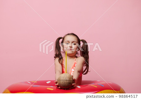 Charming 5 years old child girl holding out at camera a refreshing coconut cocktail, posing with donut swim ring over pink background 98864517