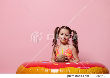 Little girl with donut swim ring, holds a coconut cocktail, cutely smiles looking at camera, isolated pink background 98864520