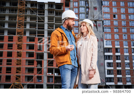 Couple in safety helmets standing outside apartment building under construction. Man and woman discussing building plan outdoors at construction site. Couple in safety helmets standing outside apartment building under construction. Man and woman discussing building plan outdoors at construction site. 98864700